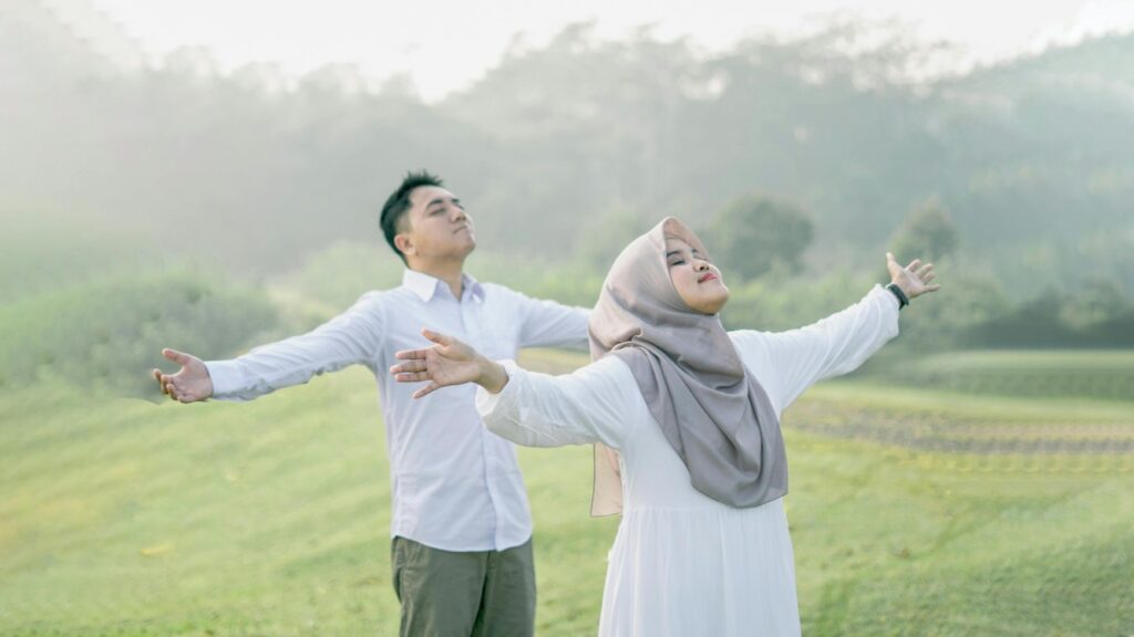 Grateful Muslim couple standing outdoors with arms spread wide, embracing thankfulness and serenity while facing the sky.